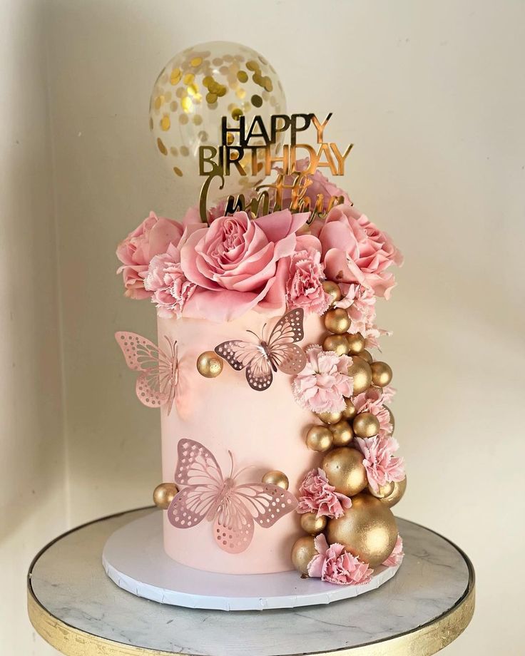 a pink and gold birthday cake decorated with flowers, butterflies and a balloon on top