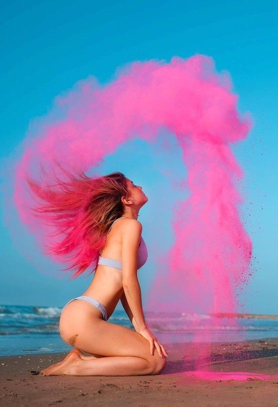 a woman sitting on the beach with pink powder coming out of her back and hair blowing in the wind