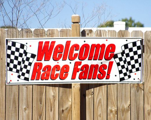 a sign that says welcome race fans on the side of a wooden fence with trees in the background