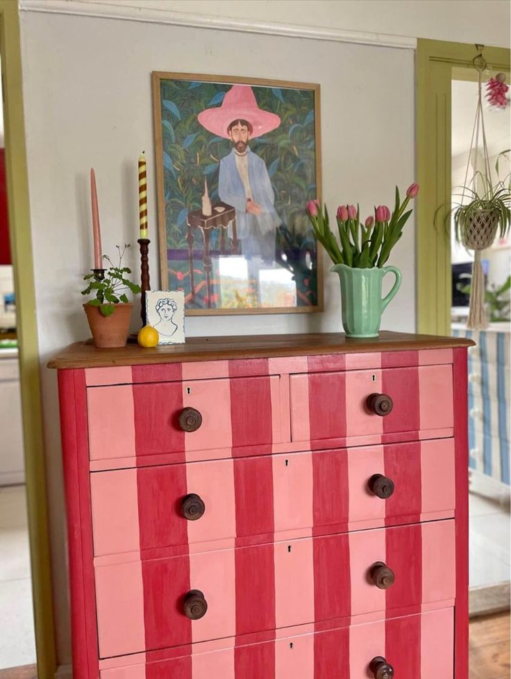 an old dresser painted pink and red with striped drawers in front of a painting on the wall