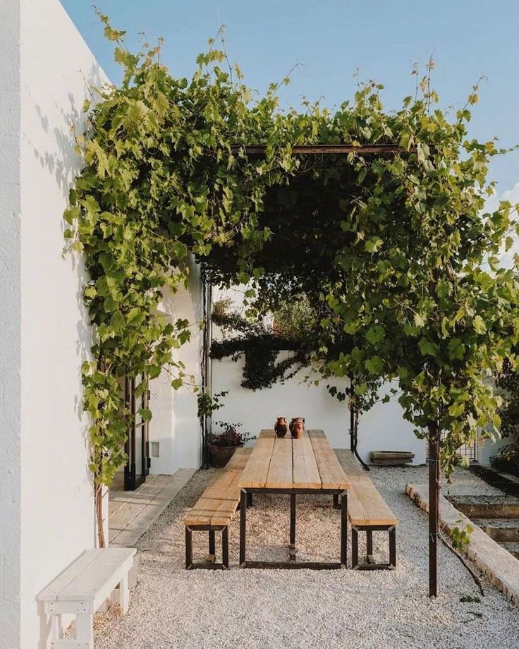 a wooden table sitting under a pergolia covered arbor in front of a white building