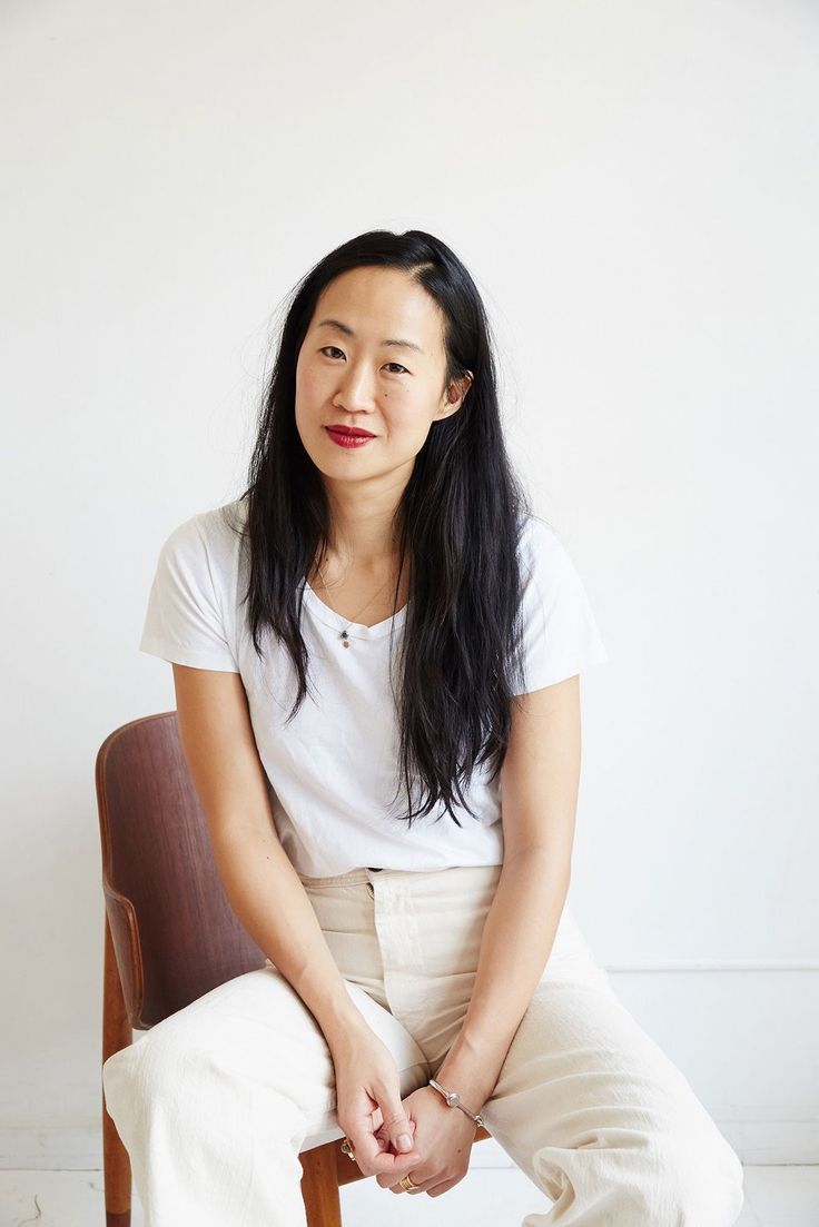 a woman sitting on top of a wooden chair in front of a white wall with her legs crossed