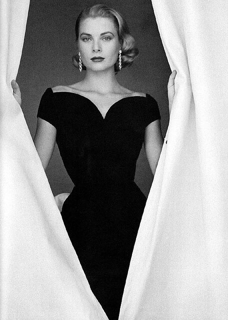 a black and white photo of a woman peeking out from behind the curtain with her hands on her hips