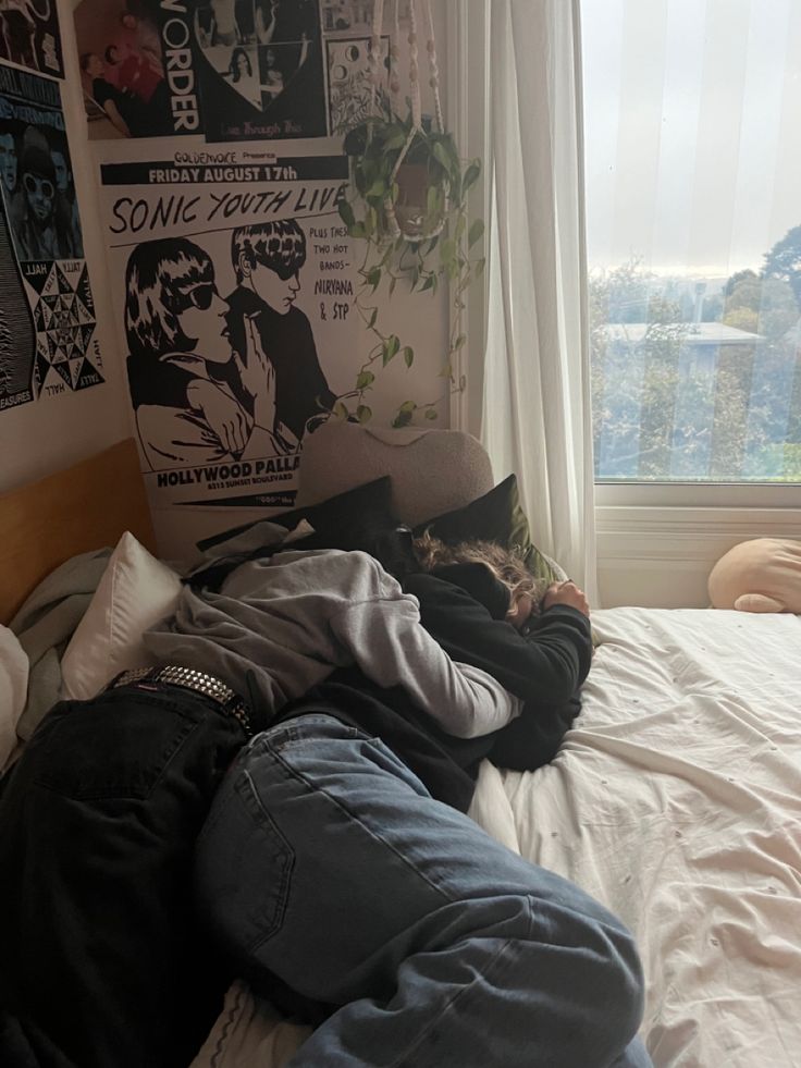 a person laying on top of a bed in front of a window next to a wall with posters