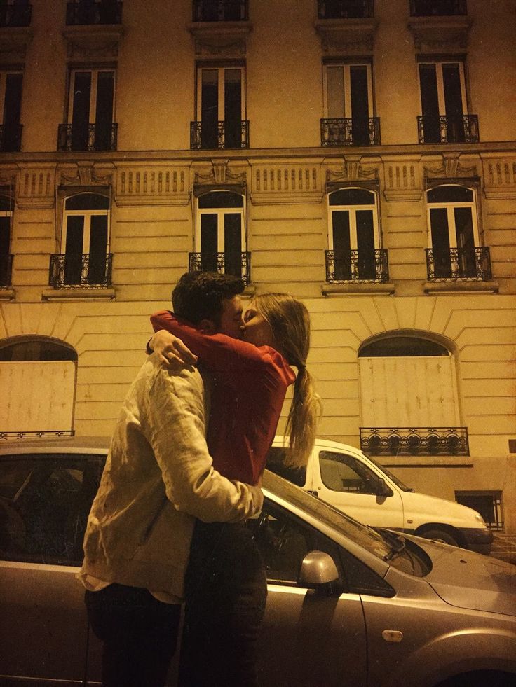 two people standing next to each other in front of a building with cars parked on the street