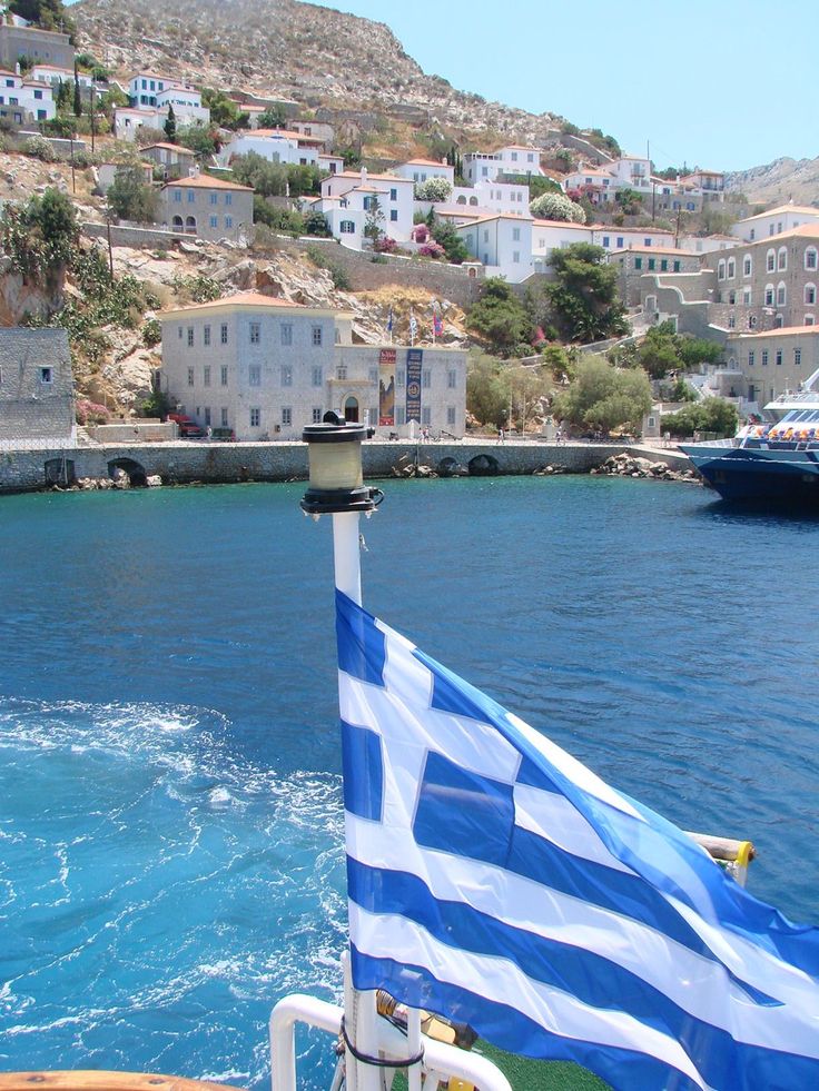 the greek flag is waving in front of some boats on the water and buildings behind it