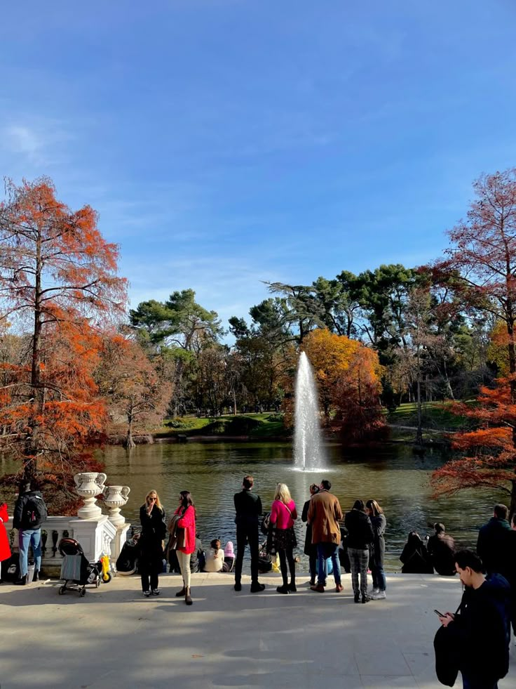 many people are standing around in front of a fountain and some trees with orange leaves