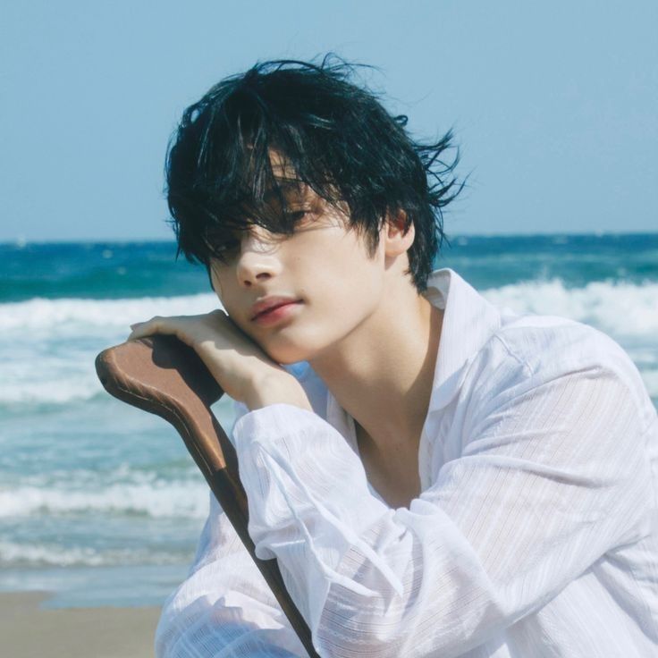 a young man sitting on top of a wooden chair near the ocean with his hand under his chin