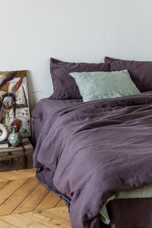 a bed with purple sheets and pillows in a room next to a painting on the wall