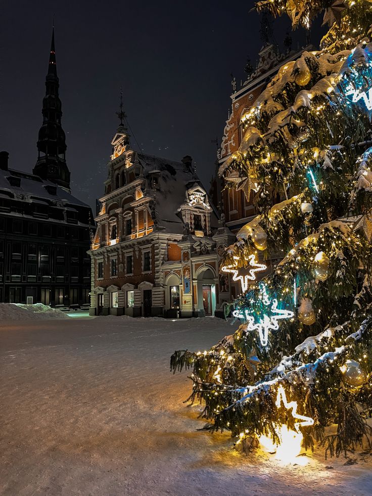 a lit up christmas tree in the middle of a snow covered area with buildings behind it