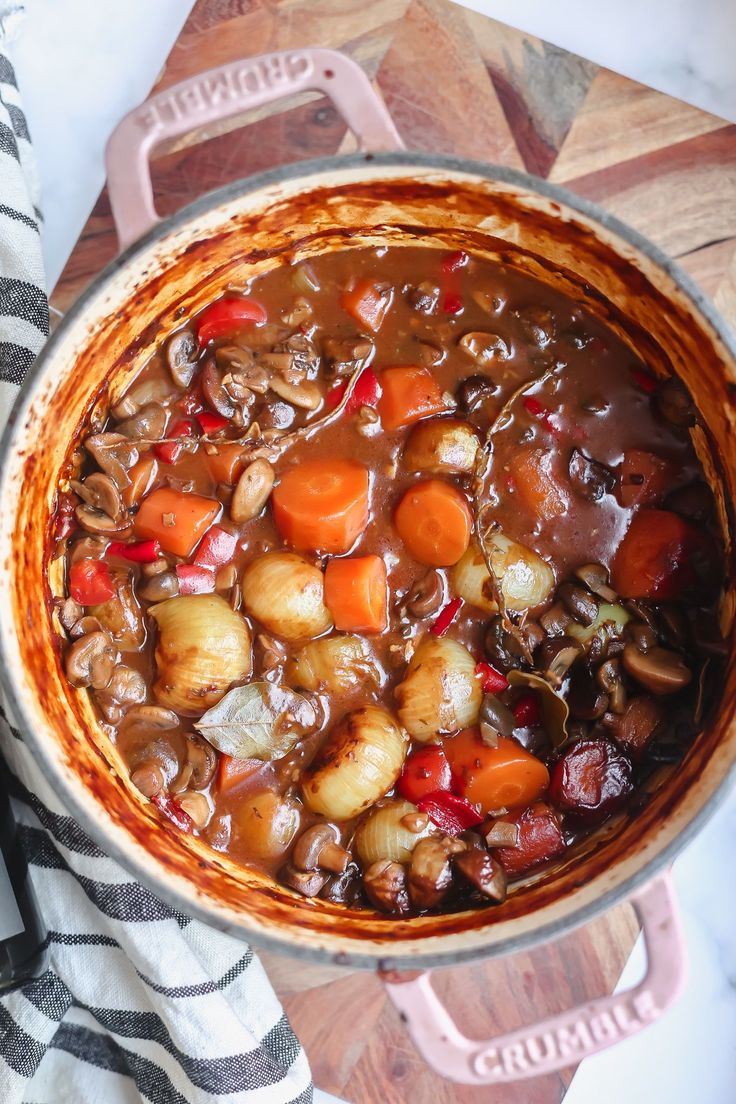 a pot filled with stew and vegetables on top of a wooden cutting board next to a towel