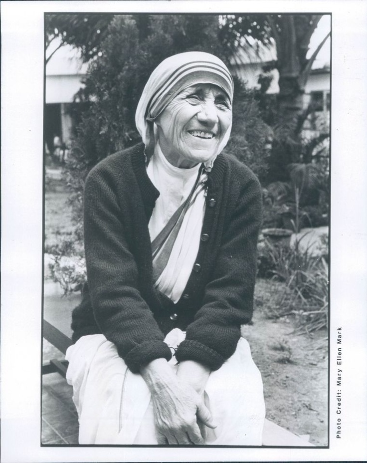 an old woman is sitting on a bench in front of some trees and bushes, smiling