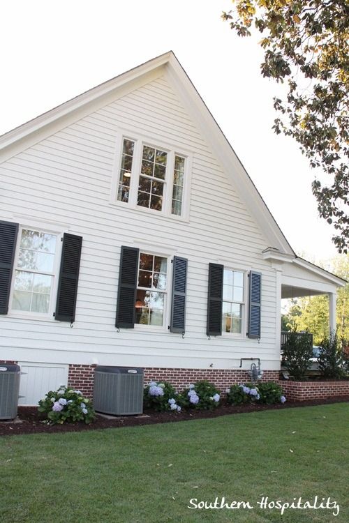 a white house with black shutters and flowers in the front yard on a sunny day