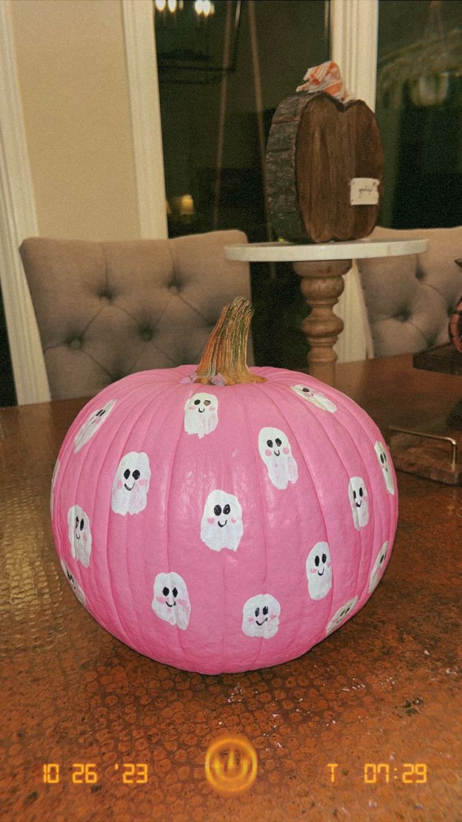 a pink pumpkin with ghost faces on it sitting on a table in front of a chair