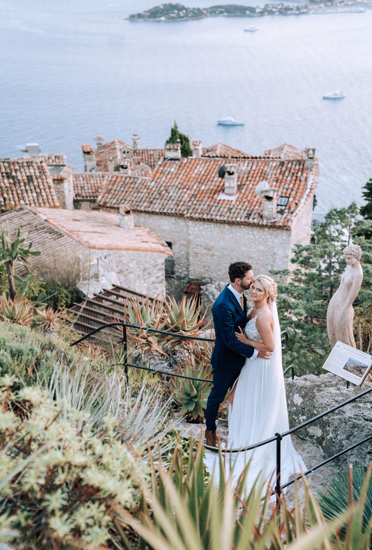 a bride and groom standing on top of a hill next to the ocean with an island in the background