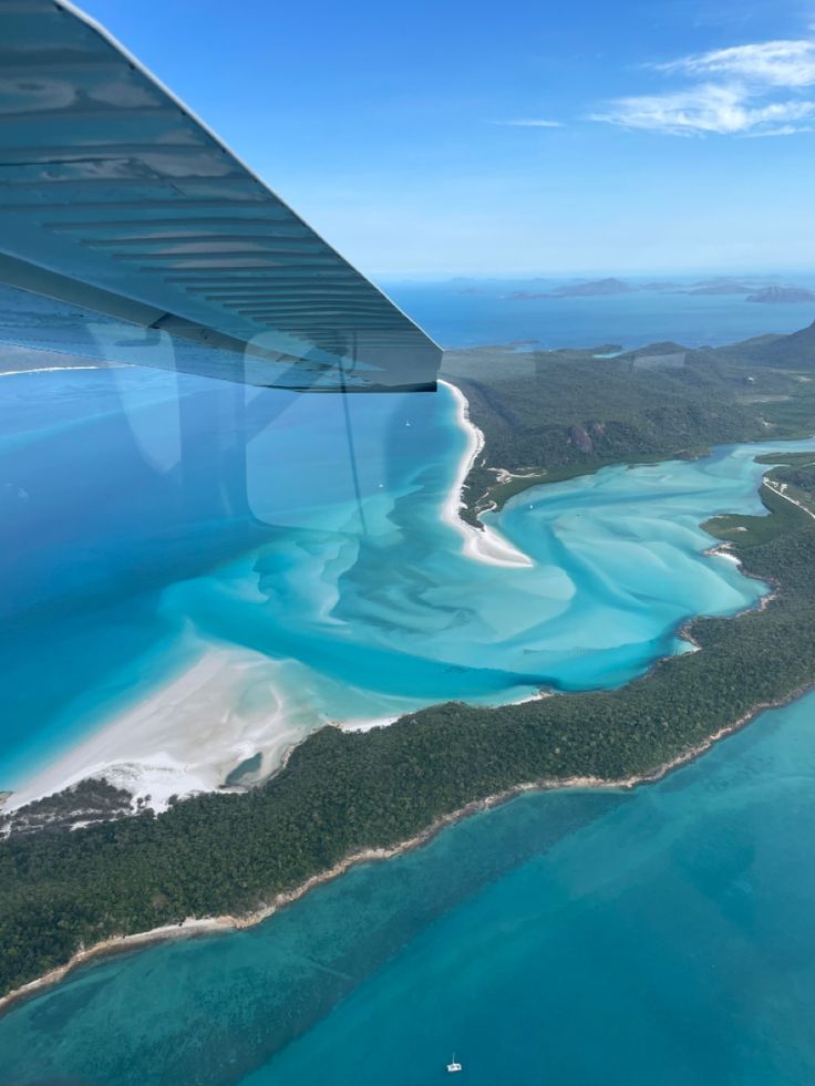 an airplane wing flying over the ocean and land near some trees in the distance with blue water