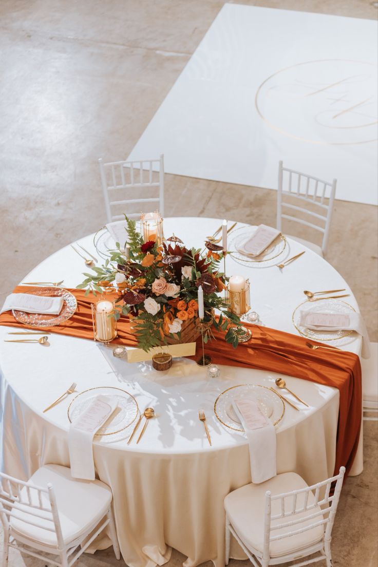 the table is set with white chairs and gold place settings, along with an orange sash