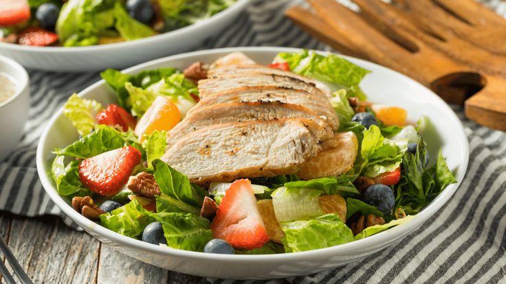 two white bowls filled with salad and fruit on top of a table next to utensils