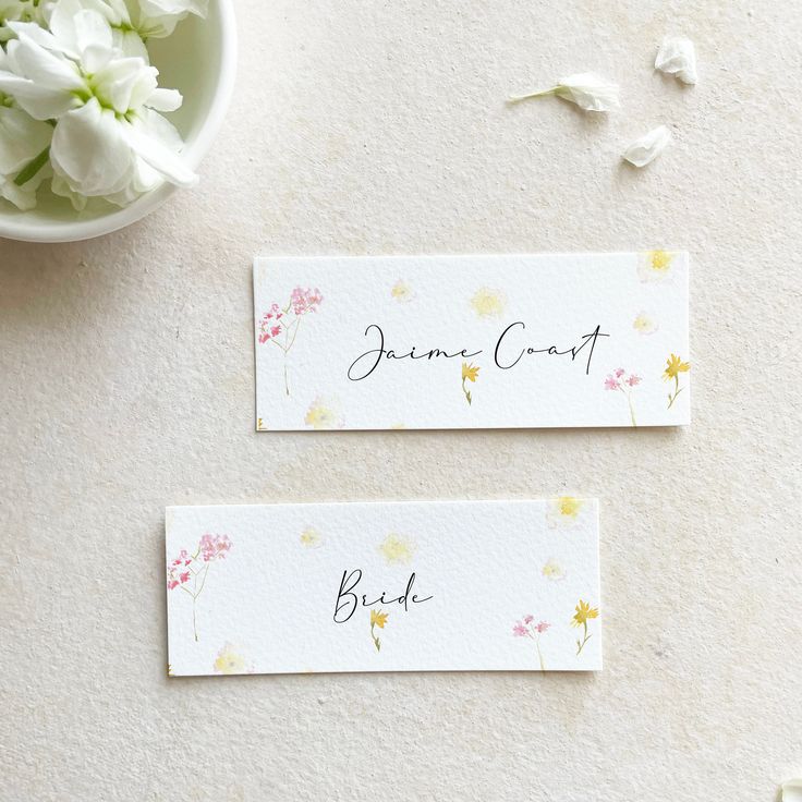 two place cards sitting on top of a table next to a bowl of white flowers