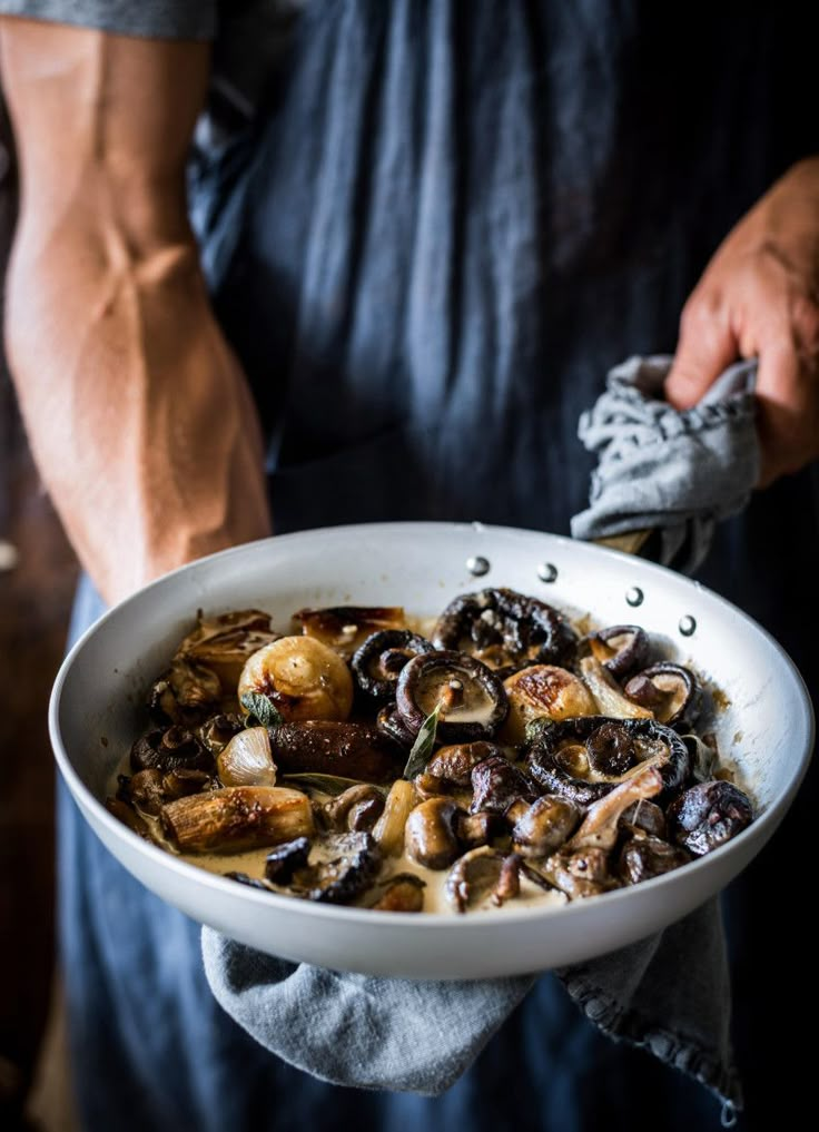 a man holding a white bowl filled with mushrooms and other food items on top of a table