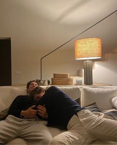 two men laying on top of a white couch next to a table with a lamp