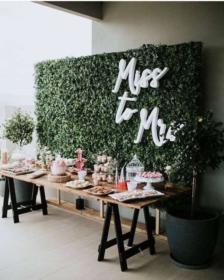 a table topped with lots of desserts next to a wall covered in greenery