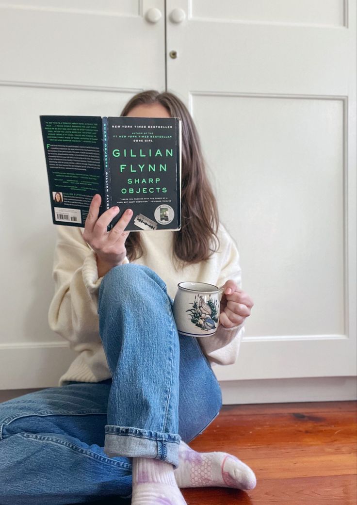 a woman sitting on the floor reading a book and holding a coffee cup in front of her face