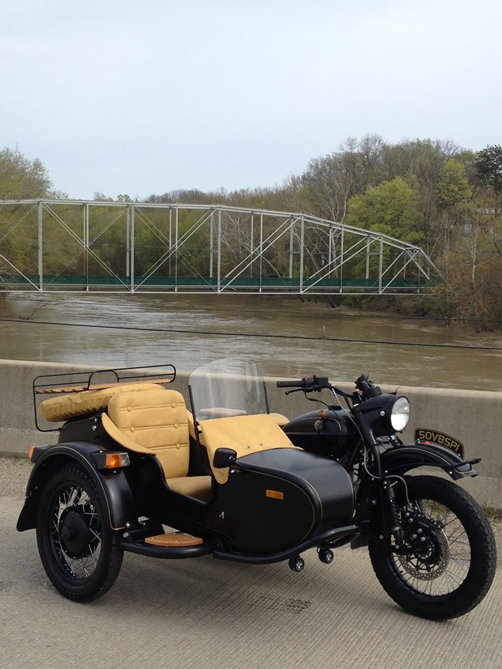 an old fashioned motorcycle parked on the side of a road near a river and bridge