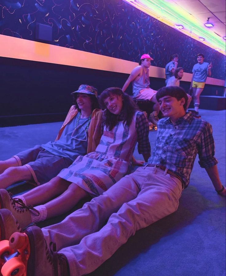 three young people sitting on the floor in front of a stage with neon lights behind them