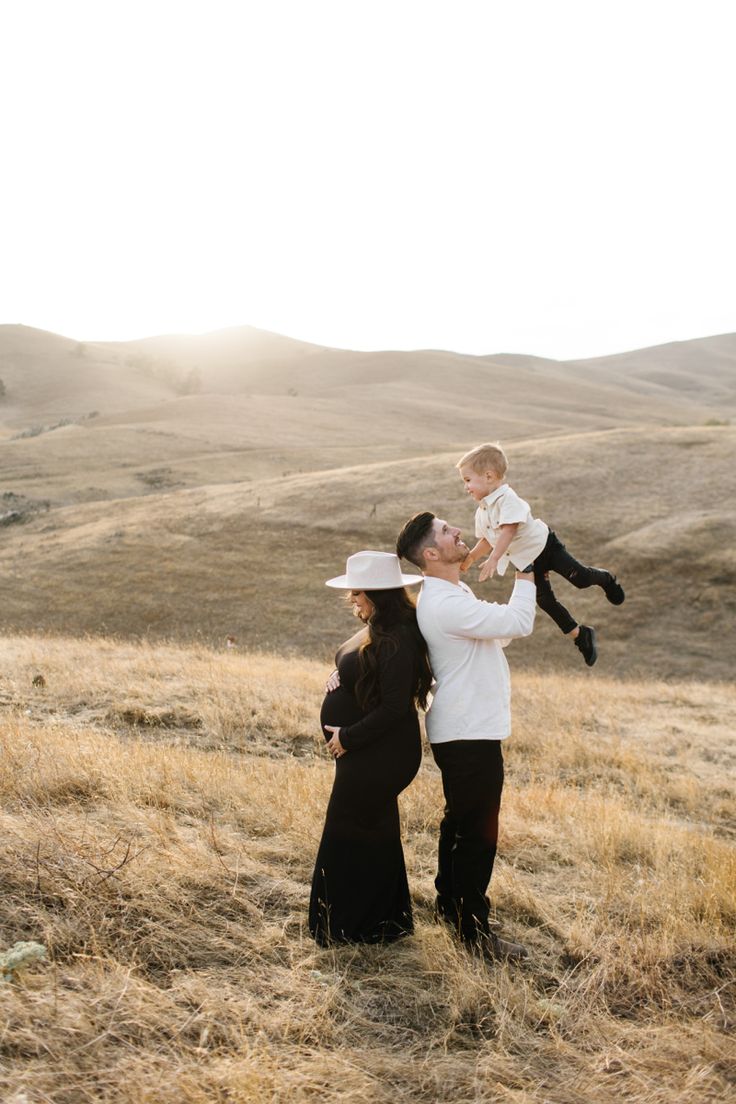 a woman holding a baby while standing on top of a grass covered field with mountains in the background