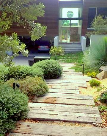 a wooden walkway in front of a house with trees and bushes on both sides, leading up to the entrance