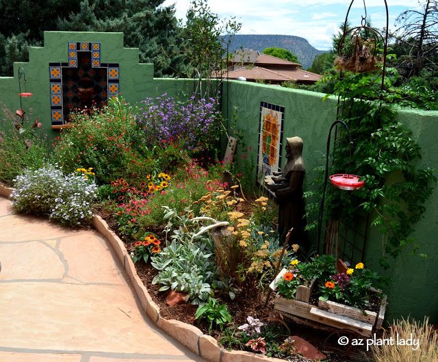 an outdoor garden with lots of flowers and plants in the center, along side a green wall