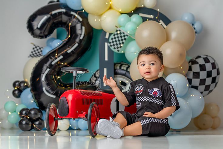 a young boy sitting on the floor in front of balloons and an old fashioned car