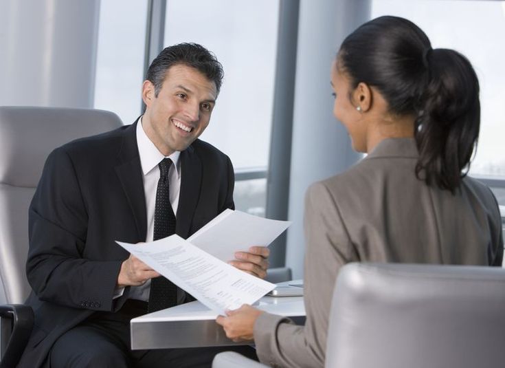 a man and woman sitting at a desk in an office talking to each other while holding papers