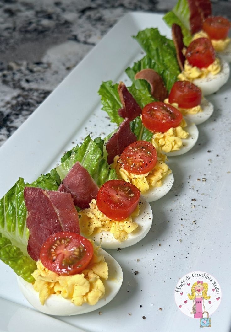 several deviled eggs with bacon, tomatoes and lettuce lined up on a white plate