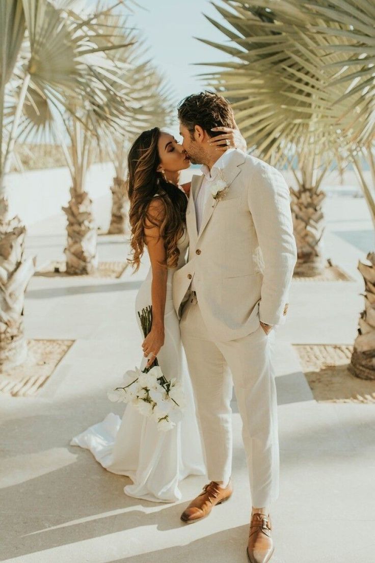 a bride and groom kissing in front of palm trees at their wedding day, wearing white tuxedos