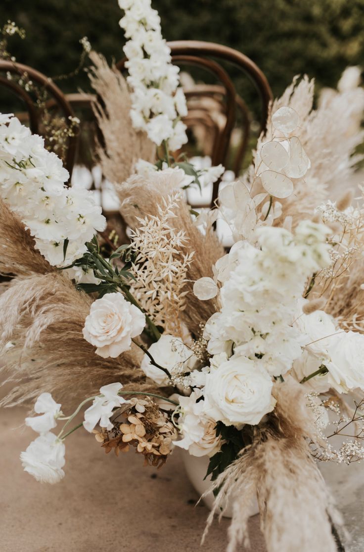 an arrangement of white flowers and grasses on a table with chairs in the back ground