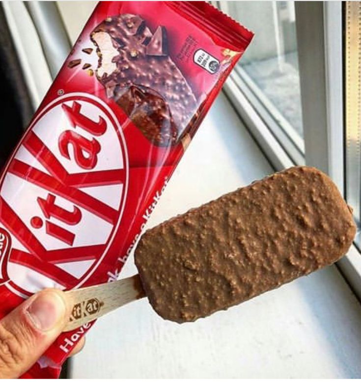 a person holding a candy bar and an ice cream sandwich on a stick in front of a window