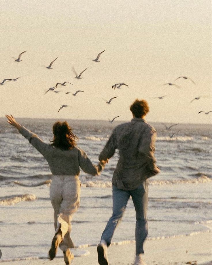 two people running on the beach with seagulls in the sky above them and one person holding hands