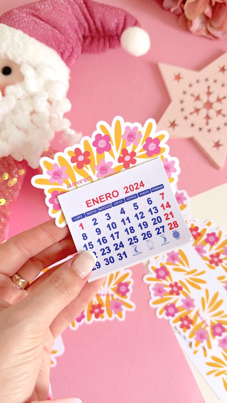 a person holding up a calendar sticker next to some flowers and other decorations on a pink surface