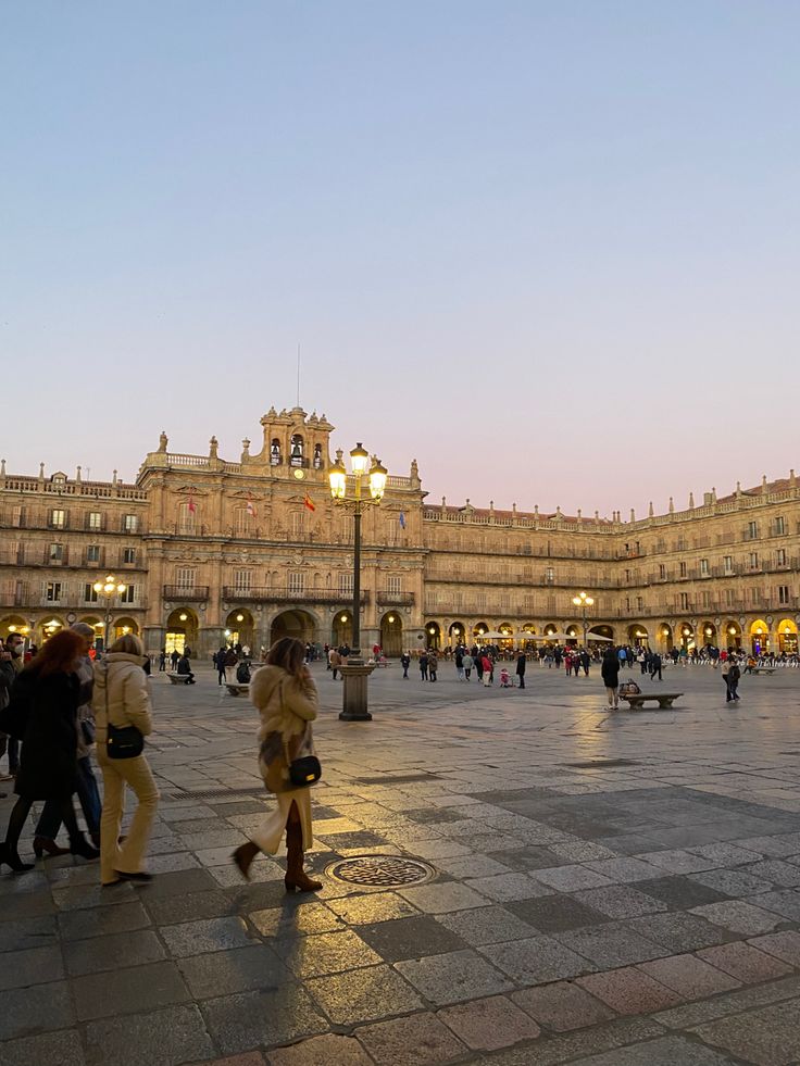 many people are walking around in an open area with buildings and lights on the sides