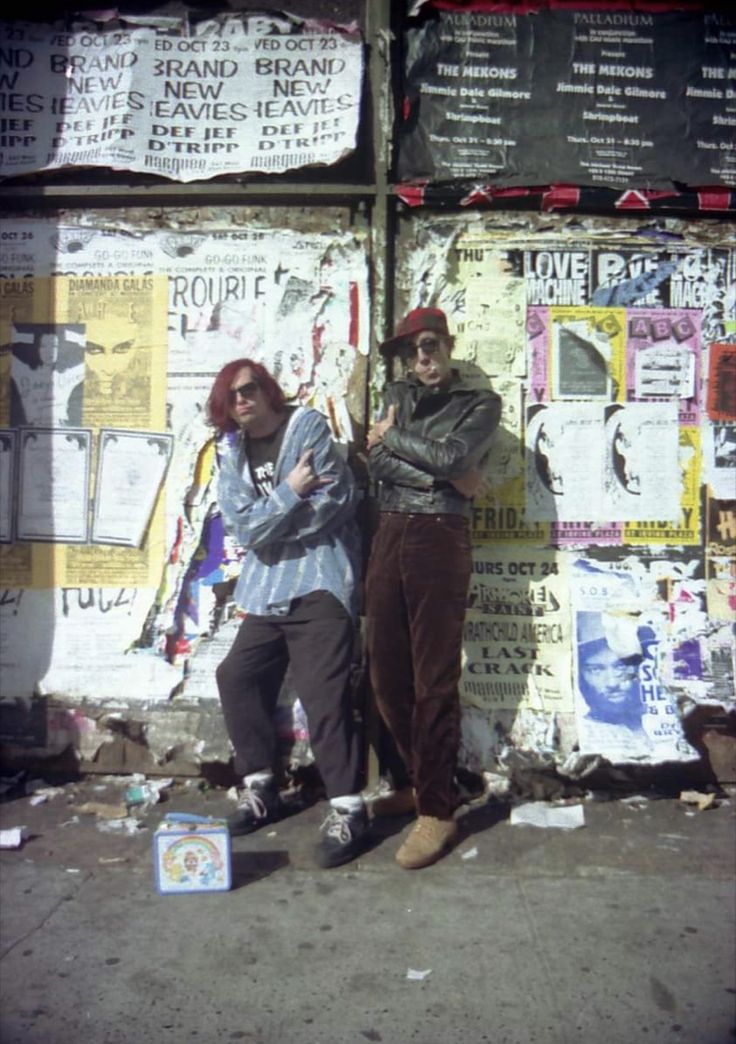 two men standing next to each other in front of a wall covered with posters and papers