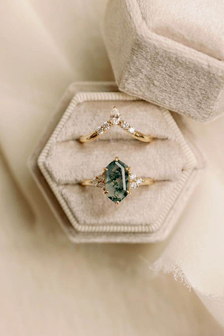 two gold rings with green and white stones on them sitting in a velvet ring box