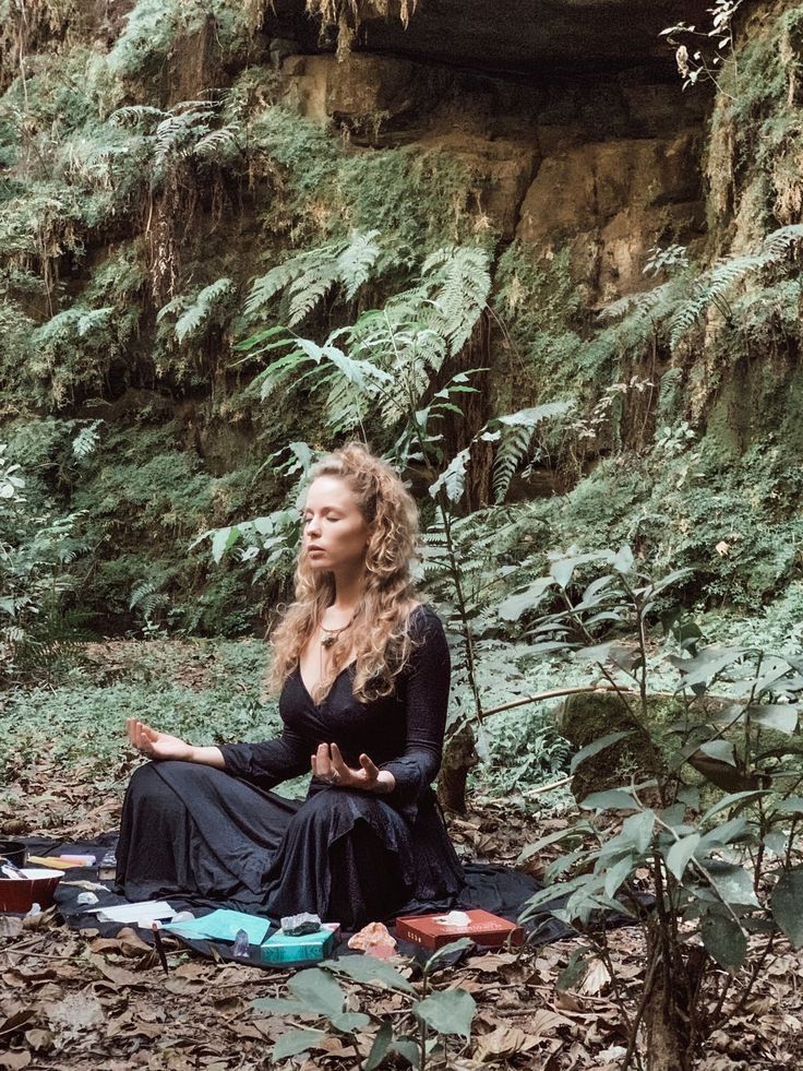 a woman sitting in the middle of a forest with her eyes closed and hands clasped