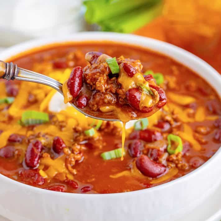 a spoon full of chili cheese soup being lifted from a white bowl with green onions and celery