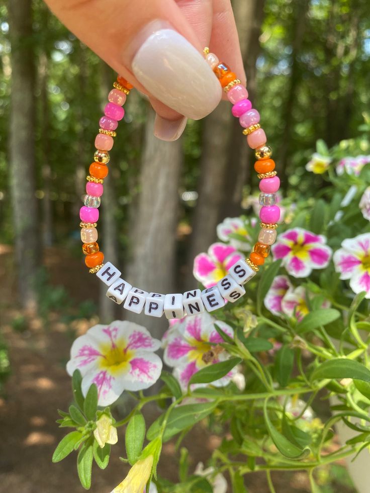 a hand holding a beaded bracelet with the word happiness written on it and flowers in the background