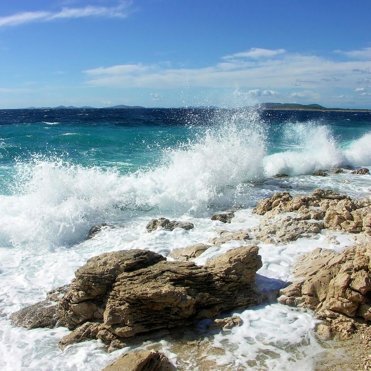 waves crashing on the rocky shore with blue sky and ocean in background, near an island