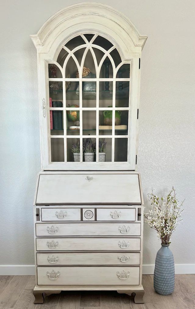 a white dresser with glass doors and drawers on it's top shelf next to a vase filled with flowers