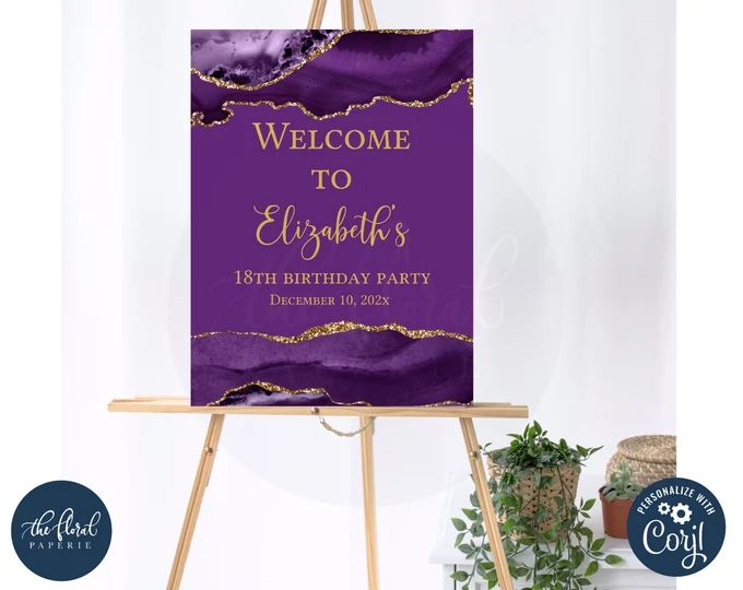 a purple and gold welcome sign sitting on an easel next to a potted plant