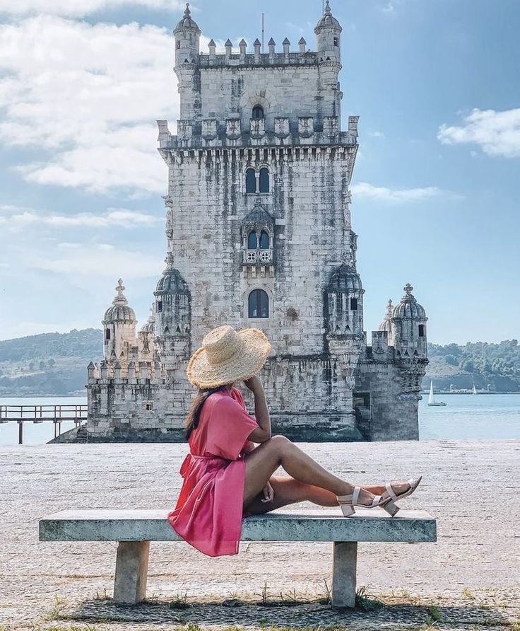 a woman in a pink dress and straw hat sitting on a bench looking at an old castle